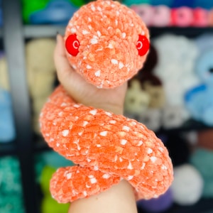 May include: A hand holding a crocheted orange and white snake with red eyes. The snake is curled up in the hand.