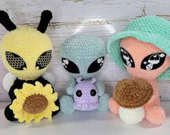 May include: Three plush alien figures. One is yellow with bee-like features and a sunflower. Another is light blue, holding a small purple creature. The third is peach-colored, wearing a mint green hat and holding a mushroom.