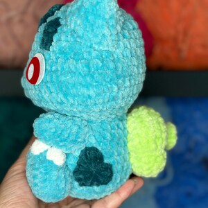 Pu&ograve; includere: Un giocattolo di peluche blu e verde all'uncinetto che ricorda un personaggio di Pokemon. Il giocattolo ha un occhio rosso e una coda verde.