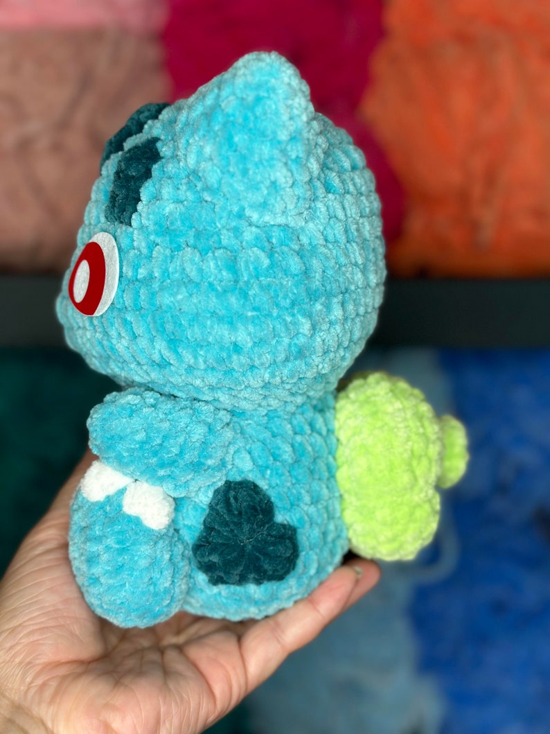Pu&ograve; includere: Un giocattolo di peluche blu e verde all'uncinetto che ricorda un personaggio di Pokemon. Il giocattolo ha un occhio rosso e una coda verde.