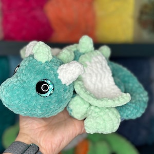May include: A crocheted green and white dragon plush toy with sparkly blue eyes. The dragon is lying on its side with its wings spread out.