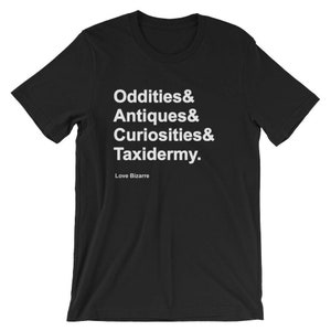 May include: Black t-shirt with white text: "Oddities & Antiques & Curiosities & Taxidermy." Below the text is "Love Bizarre." The shirt is a crew neck and short-sleeved.