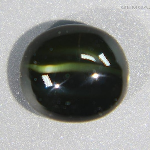 May include: A dark green, round gemstone with a prominent, light green stripe running horizontally across its center. The stone has a reflective surface with bright highlights, suggesting a polished finish. The background is a light, textured surface.