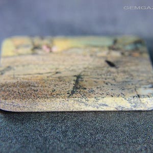 May include: A rectangular stone with rounded edges, displaying a natural pattern of beige, brown, and black tones. The stone has a textured surface and appears to be a gemstone or decorative element. The website address "GEMGAZER.COM" is visible in the top right corner.