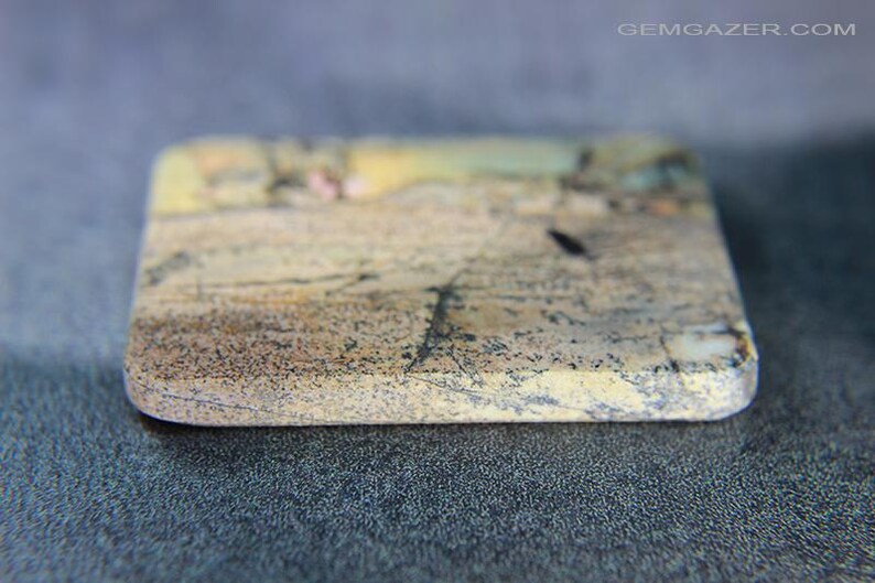 May include: A rectangular stone with rounded edges, displaying a natural pattern of beige, brown, and black tones. The stone has a textured surface and appears to be a gemstone or decorative element. The website address "GEMGAZER.COM" is visible in the top right corner.
