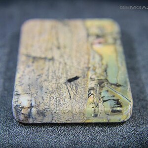 May include: A rectangular gemstone with rounded edges, displaying a mix of beige, grey, and touches of green and yellow. The stone has a natural, textured look with dark veining and markings. The surface is slightly polished, revealing the stone's unique patterns.