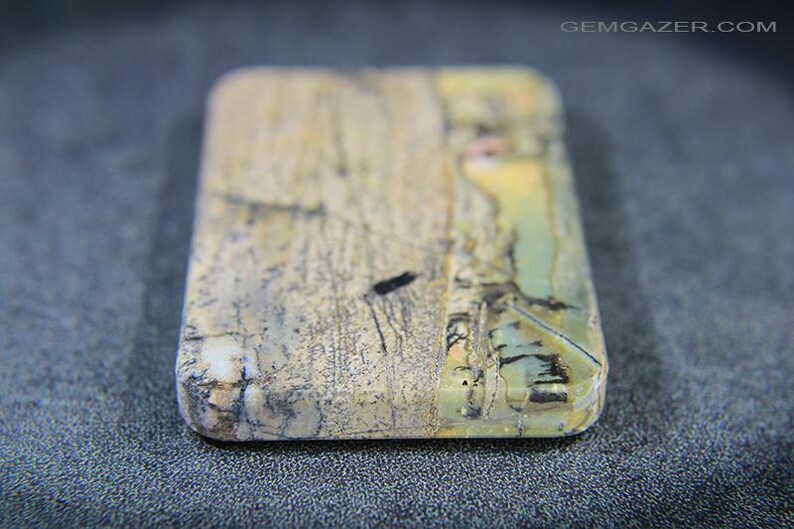 May include: A rectangular gemstone with rounded edges, displaying a mix of beige, grey, and touches of green and yellow. The stone has a natural, textured look with dark veining and markings. The surface is slightly polished, revealing the stone's unique patterns.