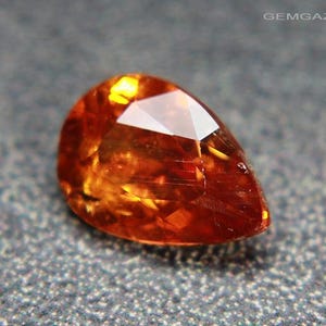 May include: A faceted, teardrop-shaped gemstone with a warm, orange-brown hue. The gem has a brilliant, reflective surface, catching the light and displaying internal facets. The gem is set against a textured, gray background.