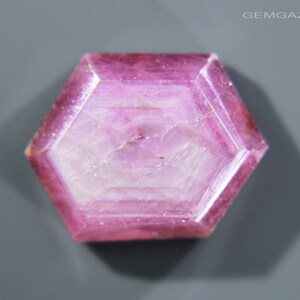 May include: A pink hexagonal ruby gemstone with a faceted cut. The gemstone has a translucent appearance with a slight shimmer.