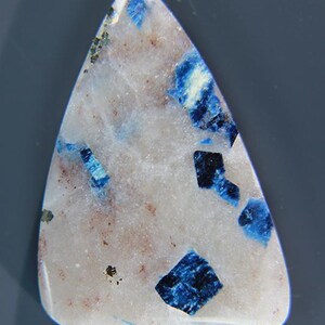 May include: A triangular gemstone with a white base, featuring striking blue and black inclusions. The stone has a polished surface and a unique pattern, with small flecks of color throughout. The image is from GEMGAZER.COM.