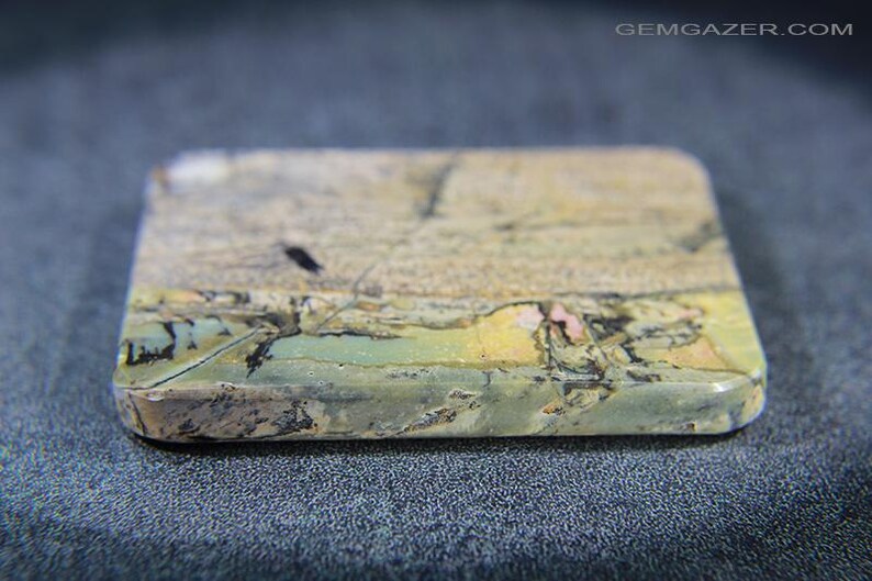May include: A rectangular gemstone with rounded edges. The stone displays a mix of colours, including beige, green, yellow, and pink, with a textured surface. The gemstone is likely used for jewellery or decorative purposes.