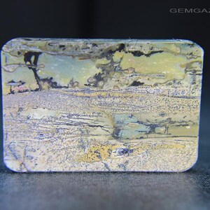 May include: A rectangular gemstone with rounded corners. The stone displays a landscape-like pattern in shades of green, yellow, grey, and black. The surface has a textured appearance, with visible lines and colour variations. The overall aesthetic is natural and earthy.