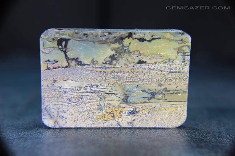 May include: A rectangular gemstone with rounded corners. The stone displays a landscape-like pattern in shades of green, yellow, grey, and black. The surface has a textured appearance, with visible lines and colour variations. The overall aesthetic is natural and earthy.