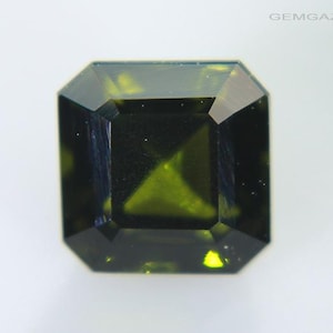 May include: A faceted, emerald-cut gemstone with a deep olive-green hue. The gem has a square shape with beveled edges, reflecting light and showcasing its internal structure. The gemstone is set against a white background.