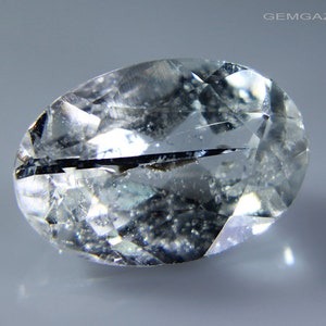 May include: A clear, oval-shaped gemstone with a black inclusion running through the centre. The gemstone has a faceted cut and is set against a grey background.