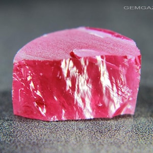 May include: A close-up of a vibrant, raw ruby gemstone. The gem is a deep, saturated pink with a rough, textured surface and a smooth, frosted top. The ruby is set against a gray, speckled background, highlighting its natural beauty.