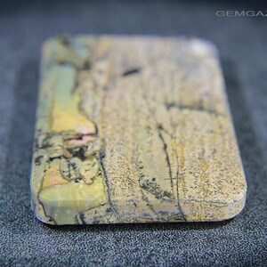 May include: A rectangular gemstone with rounded edges. The stone displays a mix of beige, yellow, pink, and black, with detailed patterns. The surface has a textured appearance, set against a dark background.