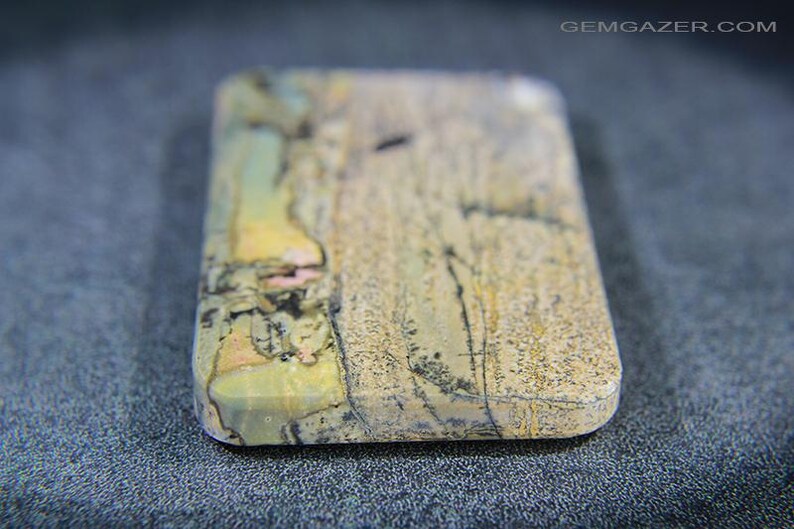 May include: A rectangular gemstone with rounded edges. The stone displays a mix of beige, yellow, pink, and black, with detailed patterns. The surface has a textured appearance, set against a dark background.