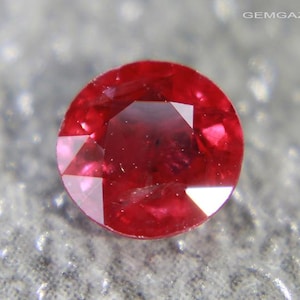 Puede incluir: Primer plano de un rubí redondo y facetado. La gema es de color rojo intenso, con múltiples facetas que reflejan la luz. El rubí se encuentra sobre un fondo gris texturizado. La imagen destaca la claridad y el brillo de la joya.