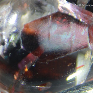 Pu&ograve; includere: Primo piano di una gemma rossa, trasparente e iridescente con una superficie ruvida e sfaccettata. La gemma &egrave; circondata da un materiale trasparente e vetroso.