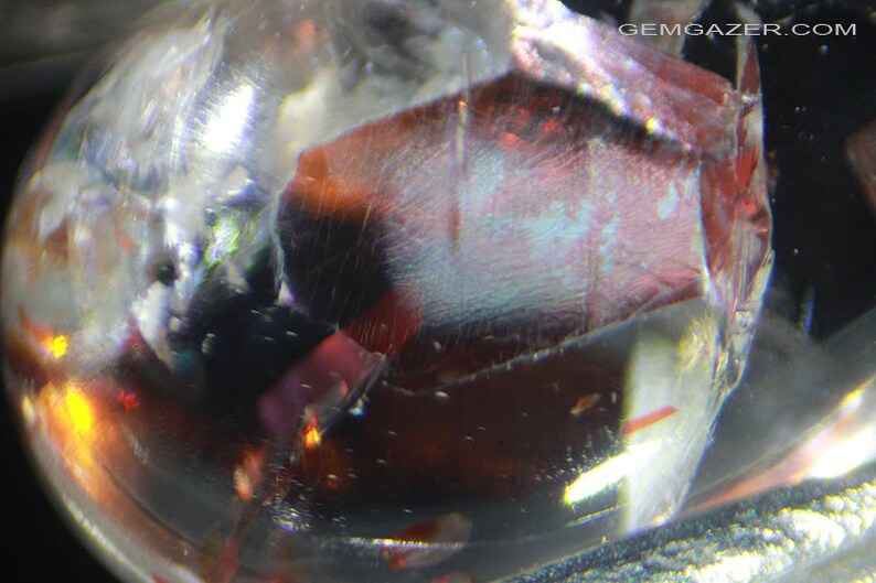 Pu&ograve; includere: Primo piano di una gemma rossa, trasparente e iridescente con una superficie ruvida e sfaccettata. La gemma &egrave; circondata da un materiale trasparente e vetroso.