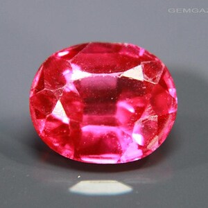 May include: A faceted, round, red gemstone with a brilliant cut. The gem reflects light, creating a sparkling effect. The gemstone is set against a grey background. The image is from GEMGAZER.COM.