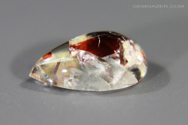 Pu&ograve; includere: Una pietra preziosa a cabochon a forma di lacrima con un corpo chiaro e traslucido e inclusiones di rosso, bianco e oro. La pietra preziosa ha una superficie liscia e lucidata.