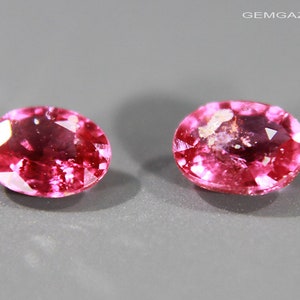 Könnte beinhalten: Zwei ovale rosafarbene Edelsteine mit leicht trübem Aussehen. Die Edelsteine liegen auf einem grauen Hintergrund.