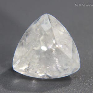 May include: A clear, triangular-cut gemstone with multiple facets. The gem is transparent, allowing light to pass through, and is set against a gray background. The website address "GEMGAZER.COM" is visible in the top right corner.
