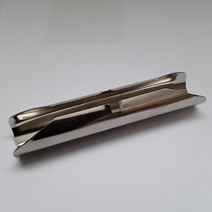 Può includere: Un posacenere in metallo argentato lucido con un design curvo a forma di canale. Il posacenere ha una superficie liscia e riflettente e una forma semplice e funzionale. I bordi sono leggermente svasati e l'aspetto generale è minimalista.