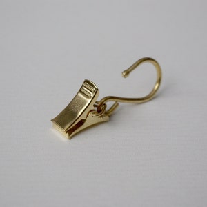 May include: A gold-toned metal clip with a hook on one end. The clip is designed to hold curtains or other lightweight items.