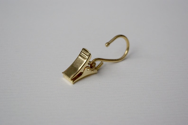 May include: A gold-toned metal clip with a hook on one end. The clip is designed to hold curtains or other lightweight items.