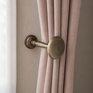 May include: A metallic curtain holdback with an antique brass finish and a circular disc design, securing a soft, dusty pink fabric curtain. The hardware features a cylindrical stem attached to a circular wall-mounted base plate.