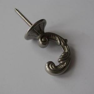Op de afbeelding: Zilveren metalen haak met een decoratief scrollontwerp. De haak heeft een schroef aan de achterkant om te bevestigen.