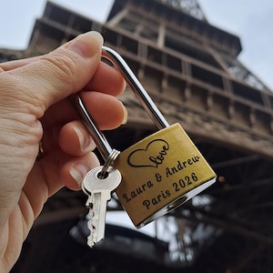 May include: A gold-toned metal padlock with a silver shackle and a single silver key attached. The padlock features a black engraved heart icon with the word love inside, followed by the text Laura & Andrew and the date Paris 2026.