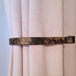 Op de afbeelding: Een donkerbruine houten gordijnband met een verweerde afwerking. De band is gebogen en heeft een afgerond uiteinde.