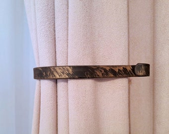 May include: A decorative, dark brown curtain tieback with a textured, wood-like appearance. The tieback is holding a light pink curtain in place. The tieback has a rectangular shape with rounded edges.