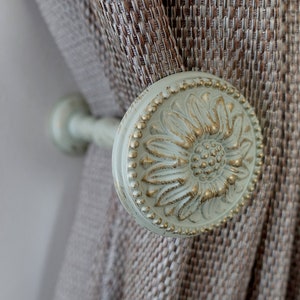May include: A close-up of a decorative curtain tieback with a floral design. The tieback is made of white metal with gold accents and features a raised flower pattern.