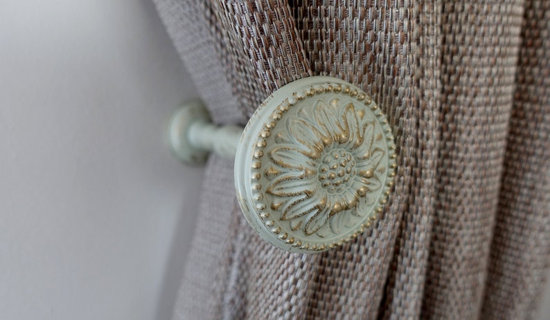 May include: A close-up of a decorative curtain tieback with a floral design. The tieback is made of white metal with gold accents and features a raised flower pattern.