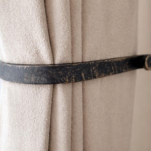 May include: A dark, distressed metal curtain tieback encircles a beige curtain. The tieback has a flat, rectangular shape with rounded ends and a textured, aged appearance, adding a rustic touch to the window treatment.