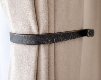May include: A dark, distressed metal curtain tieback encircles a beige curtain. The tieback has a flat, rectangular shape with rounded ends and a textured, aged appearance, adding a rustic touch to the window treatment.