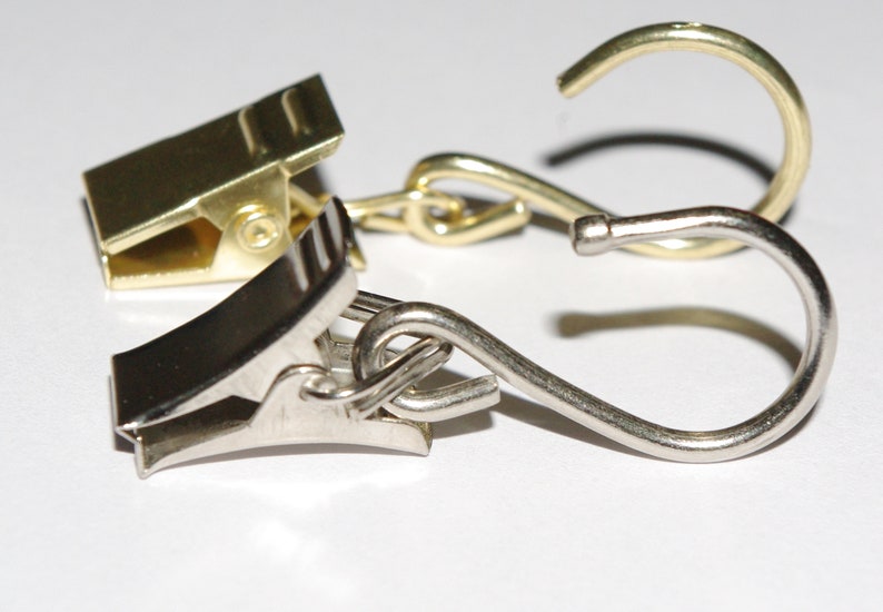 May include: Two metal curtain clips, one gold and one silver, connected by a chain. The silver clip has a hook on the end.