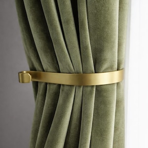 May include: Olive green velvet curtains secured by a gold-coloured metal curtain tieback. The tieback is a simple band with a small, rectangular end piece. The curtains are draped in soft folds.