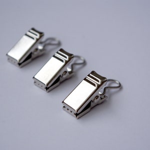 Puede incluir: Tres clips metálicos plateados y brillantes dispuestos en diagonal sobre una superficie blanca. Cada clip tiene un cuerpo rectangular con un pequeño patrón de agujeros y un lazo de alambre curvado.