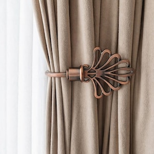 May include: A decorative bronze curtain tieback with a stylized floral design holds a beige curtain. The curtain is made of a soft, textured fabric. White sheer curtains are visible in the background.