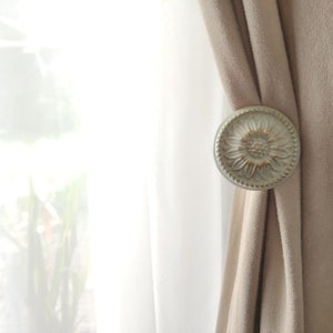 May include: A silver curtain tieback with a floral design. The tieback is holding back a pair of beige curtains.