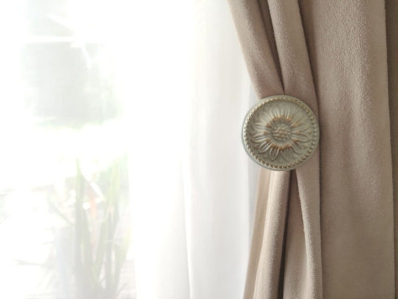 May include: A silver curtain tieback with a floral design. The tieback is holding back a pair of beige curtains.