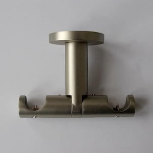 Może przedstawiać: Uchwyt do drążka na zasłony ze szczotkowanego niklu. Uchwyt ma okrągłą podstawę i pionowy słupek, który łączy się z poziomym prętem z dwoma zakrzywionymi końcami. Sprzęt jest przeznaczony do trzymania drążka na zasłony. Wykończenie jest matowe srebrne.