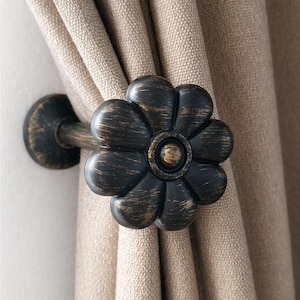 Pode incluir: Um prendedor de cortina de cor bronze com um design em forma de flor. O prendedor é feito de metal e tem um acabamento escuro e envelhecido. A flor tem pétalas arredondadas e um centro circular. Segura uma cortina bege.