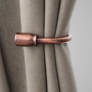 May include: A copper-colored curtain tieback holds a section of a light gray curtain. The tieback has a cylindrical end piece and a curved, rod-like design. The curtain fabric has a textured appearance.